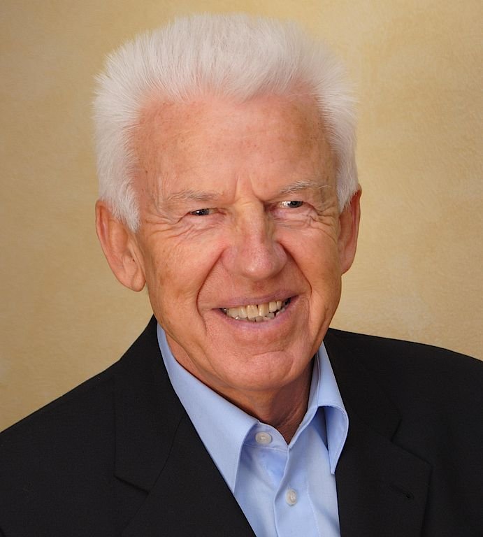 Der Mann hat ein strahlendes Lächeln und trägt einen schwarzen Blazer über einem hellblauen Hemd, vor einem sanften Hintergrund.