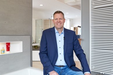 Professioneller Mann in modernem Badezimmer-Design Ein Mann in einem blauen Anzug sitzt entspannt in einem modernen Badezimmer mit eleganten Fliesen und einem großen Spiegel.